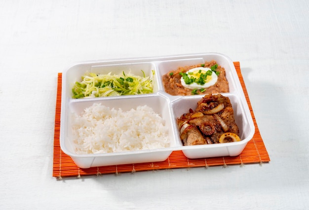 Food delivery, styrofoam lunch box with fried steak with onion, zucchini, rice, beans and boiled egg on white wood texture.