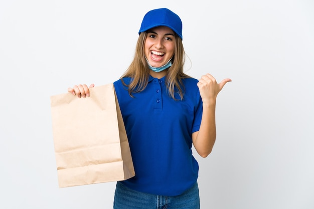 Food delivery woman and protecting from the coronavirus with a mask over isolated white wall pointing to the side to present a product