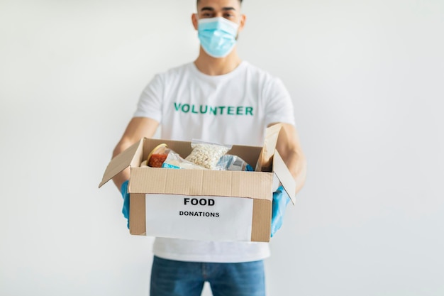 Food donations Millennial arab man in volunteer tshirt and medical mask showing box with canned food and packages