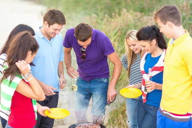 food, eating, cooking, summer holidays and people concept - group of friends having picnic and making barbecue on beach