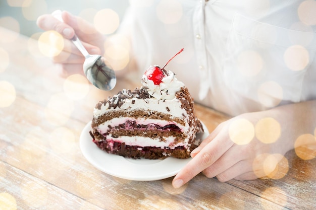 food, junk-food, culinary, baking and holidays concept - close up of woman eating chocolate cherry cake with spoon and sitting at wooden table over holidays lights background