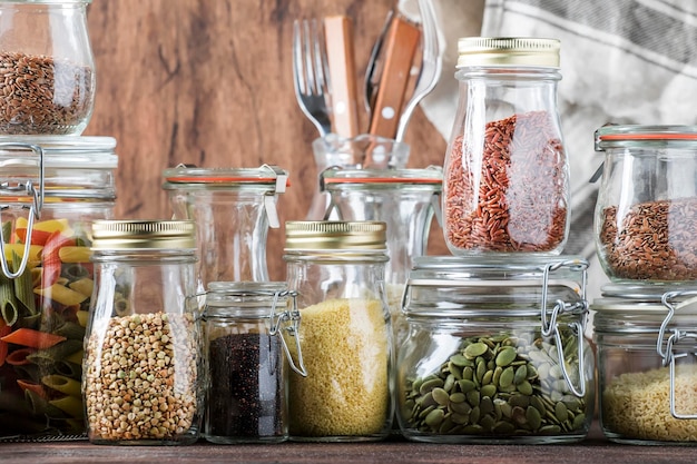 Food set Raw cereals pasta groats organic legumes and useful seeds in glass jars Vegan source of protein and energy resources Rustic wooden kitchen table background