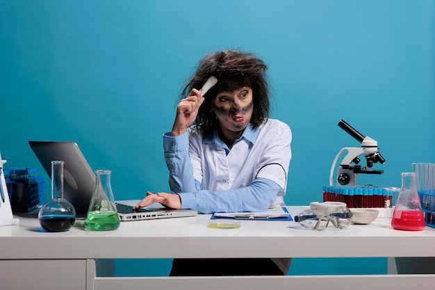 Foolish goofy biochemistry expert scratching head while trying to understand why chemical experiment led to explosion. Confused lunatic scientist with wild look sitting at desk in laboratory.