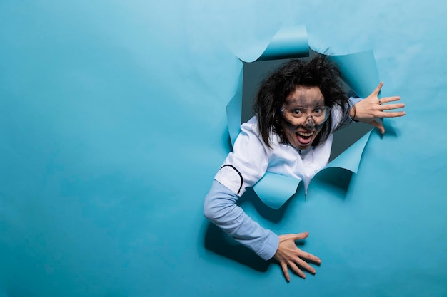 Foolish humorous scientist with comic expression on blue background. Young adult chemist with wacky look, messy hair and dirty face acting goofy after experiment explosion. Studio shot.