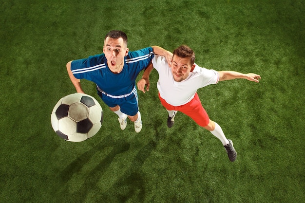 Football players tackling for ball over green grass background. Professional football soccer players in motion at studio. Fit jumping men in action, jump, movement at game. Top view