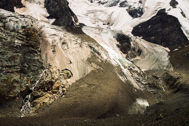 Foothills of giant glacier. Amazing rocky relief with snow and ice. Wonderful huge mountain rocky natural wall with small waterfalls. Water from glacier. Fantastic artwork of majestic highland nature.