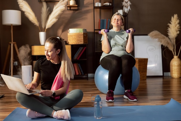 In foregroung happy young girl sitting on matt holding laptop in hands searching exercises