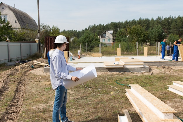 Foreman checking building supplies on site standing in front of a stack of lumber and timber with a blueprint in his hands watching workmen working