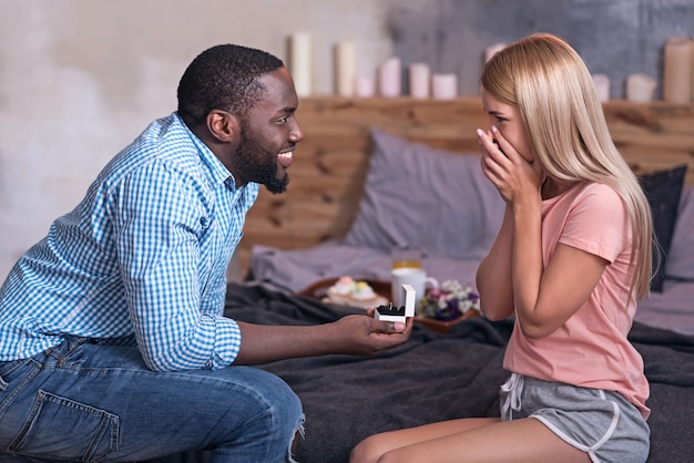 Forever and ever. African young smiling man giving a wedding ring and making a proposal to his pretty girlfriend while she feeling amused.