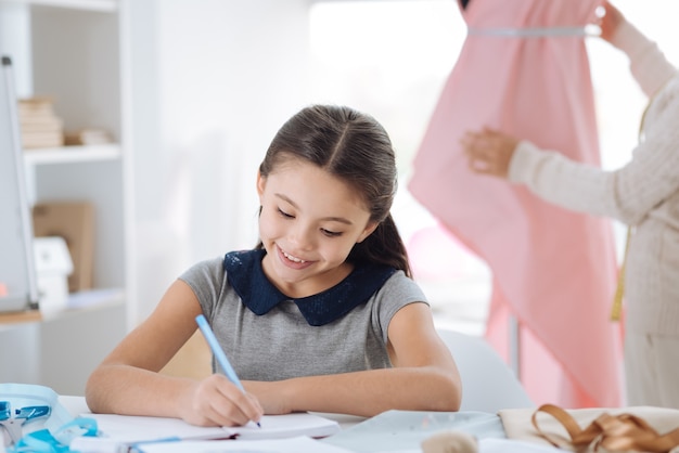 Not to forget. Smart positive creative girl sitting at the table and writing in her notebook while noting down her creative ideas