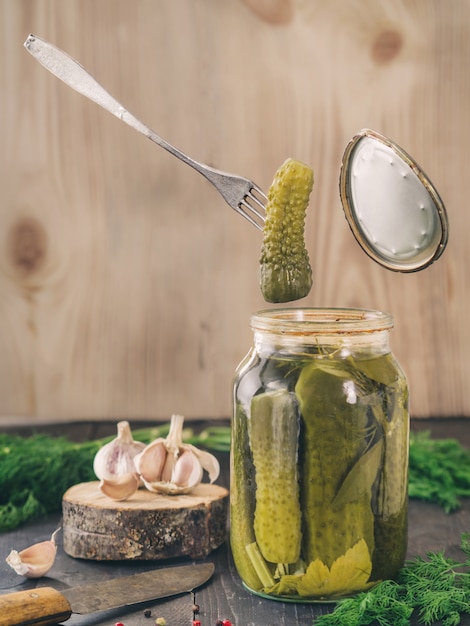 The fork holds a pickled cucumber that just has been taken from glass jar pickled cucumbers on wooden background pickles in glass jar with garlic peppercorn and fresh dill flying food