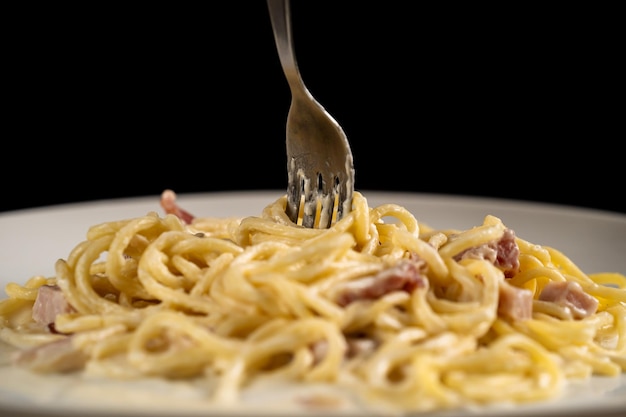 Fork is spinning carbonara spaghetti on white plate on black background