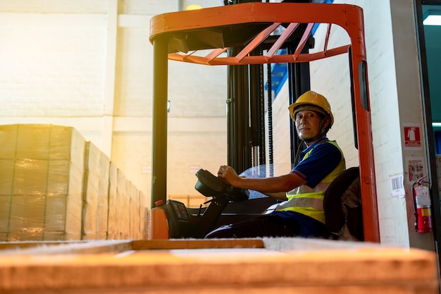 Forklift driver asian elderly man in safety jumpsuit uniform with yellow hardhat at warehouse. Worker male senior looking at camera and smiling in forklift loader work