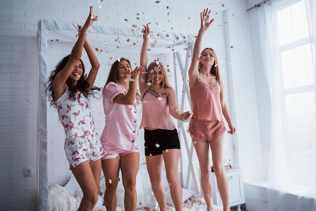 Photo four attractive women in pajamas smiling and gesturing confetti in the air young girls have fun on the white bed in nice room