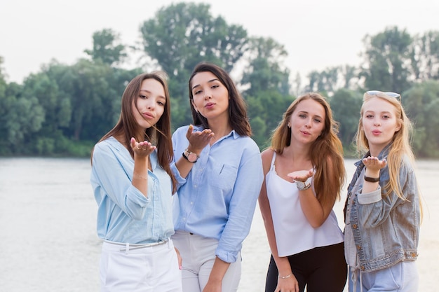 Four attractive young women standing and sending kisses at camera outdoor. trees and river at background