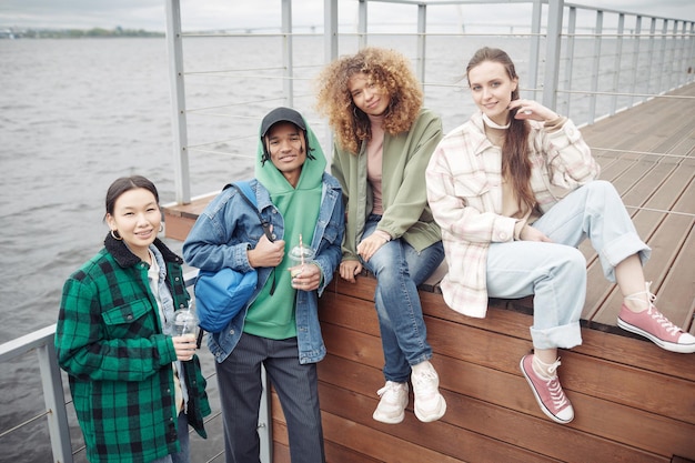 Photo four happy teenagers in casualwear resting on wooden pier on weekend