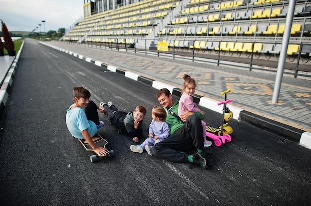 Four kids with father in asphalt play and having fun. Sports family spend free time outdoors with scooters and skates.