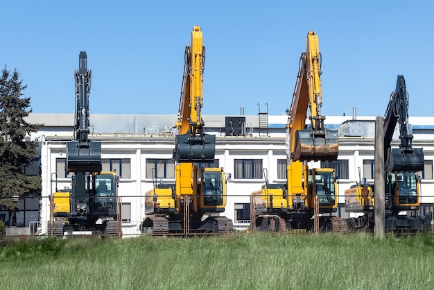 Four new modern yellow and black excavators stand behind a metal fence against a twostory building Diggers with a buckets on sale