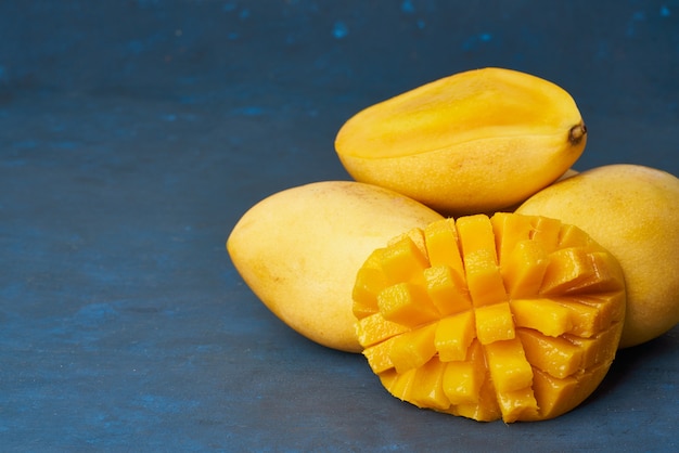 Four whole mango fruits on dark blue table and cut into slices. Large juicy ripe yellow fruits