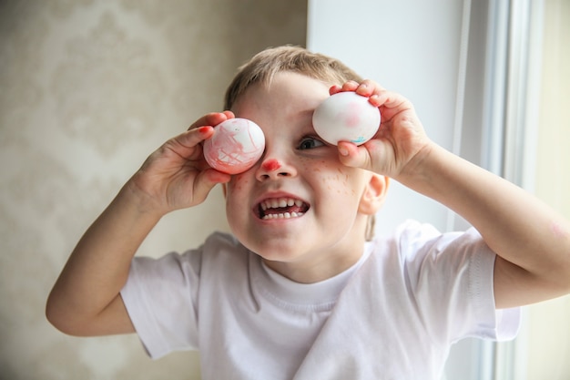 A four-year-old boy in a white T-shirt and a painted nose is fooling around trying on Easter eggs on his eyes