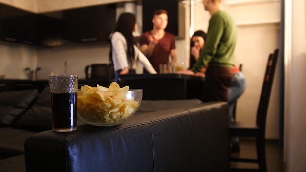 Four young people standing on kitchen and talking bowl filled with chips in focus on the foreground