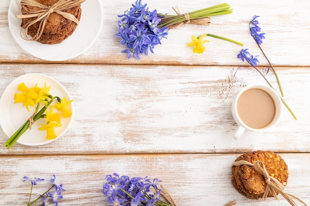 Frame composition with oatmeal cookies spring snowdrop flowers bluebells narcissus and cup of coffee on white wooden background top view copy space