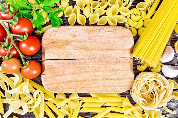 Frame from a wooden plate, different pasta, tomatoes, garlic and parsley on a wooden board background