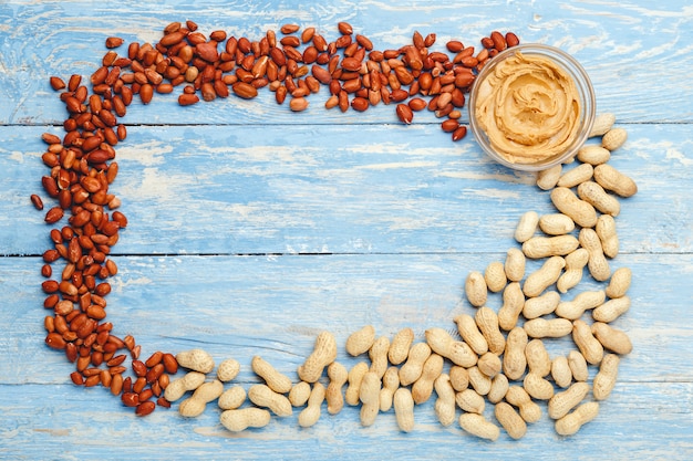 Frame made of nuts peanuts in shell, peeled peanuts, peanut butter in glass plate with copy space in the center on blue wooden background. Flat lay with place for text for peanut butter, peanut paste