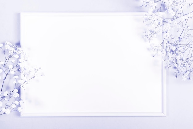 Frame with white copy space and gentle gypsophila flowers around. Color of the year 2022 Very Peri. Trendy creative holiday backdrop. View from above.