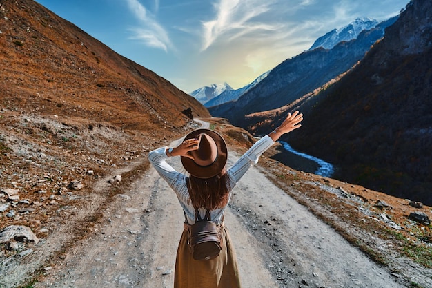 Free hipster girl wanderer with open arms wearing backpack and hat walking alone on a mountain path, enjoying freedom and happy travelling. Go travel