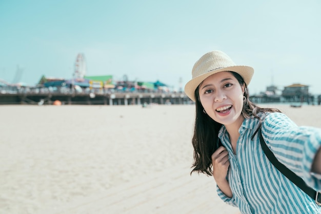 freedom young asian japanese female backpacker wear straw hat standing on beach with blue sky. woman photographing selfie with amusement park and sea on sunny day. lady face camera make self portrait