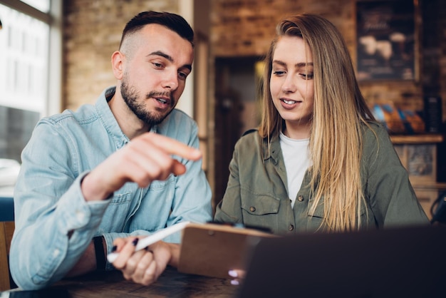 Freelance adult female with clipboard working with adult male surfing laptop in cafe