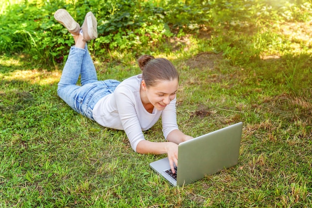 Freelance business concept. Young woman lying on green grass lawn in city park working on laptop pc computer. Lifestyle authentic candid student girl studying outdoors. Mobile Office