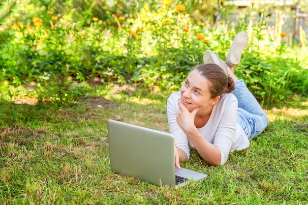 Freelance business concept. Young woman lying on green grass lawn in city park working on laptop pc computer. Lifestyle authentic candid student girl studying outdoors. Mobile Office