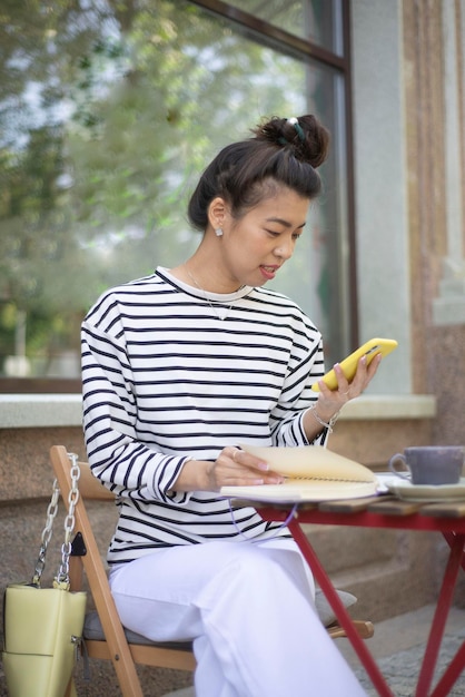 A freelance girl works with mobile phone writing in notebook in an outdoor cafe