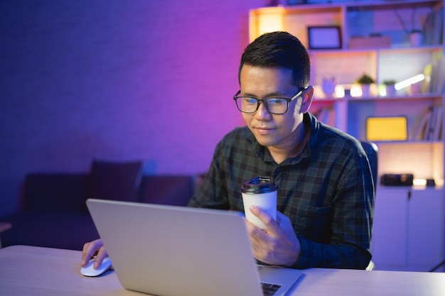 Freelance man working on laptop and holding cup of hot coffee at home at night Man freelancing smiling relaxing after finishing work at home at night Freelance work and work from home concept