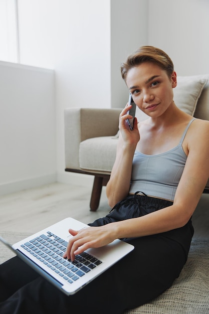 Freelance woman with laptop and phone works from home sitting on the floor in her home clothes with a short haircut free copy space