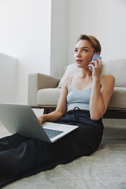 Freelance woman with laptop and phone works from home sitting on the floor in her home clothes with a short haircut free copy space