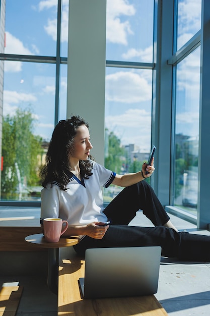 Freelance work A young smiling curlyhaired woman sits near the large windows in a cafe and works on a laptop Remote work A woman is talking on the phone while sitting in a cafe