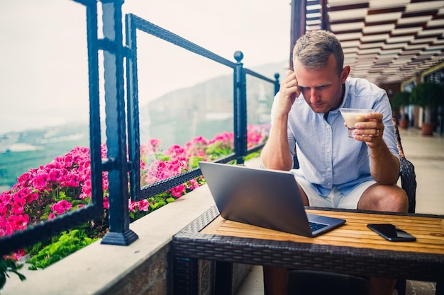 Freelancer concept Successful young man businessman working on a laptop computer sitting on the terrace drinking coffee and talking on the phone