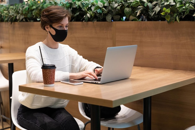 Freelancer girl in a shopping center at the food court works behind a laptop in a mask from covid