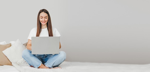 A freelancer girl sits on a white bed with a laptop looks at the monitor and works