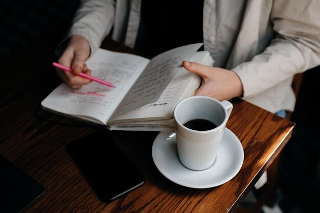 A freelancer girl works in a cafe drinks cold coffee and makes notes in a notebook with a pink pen