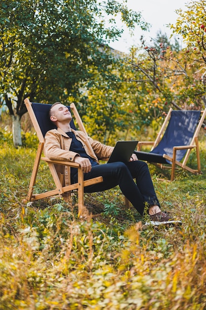 Freelancer man with laptop working in garden sitting in wooden chair outdoors A young blogger works on a computer in a public park Place of work remote work