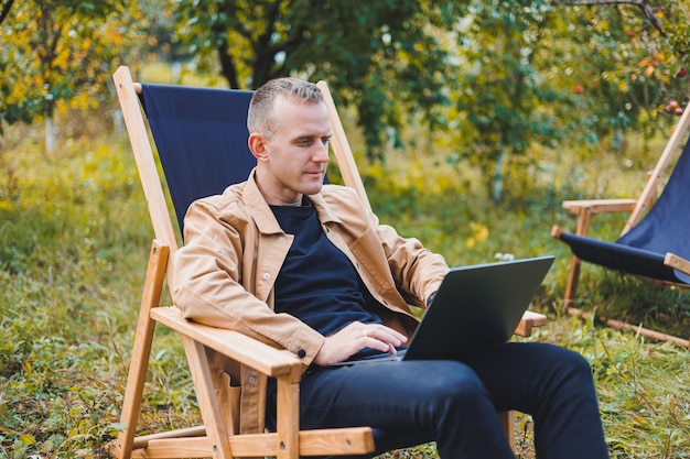 Freelancer man with laptop working in garden sitting in wooden chair outdoors A young blogger works on a computer in a public park Place of work remote work