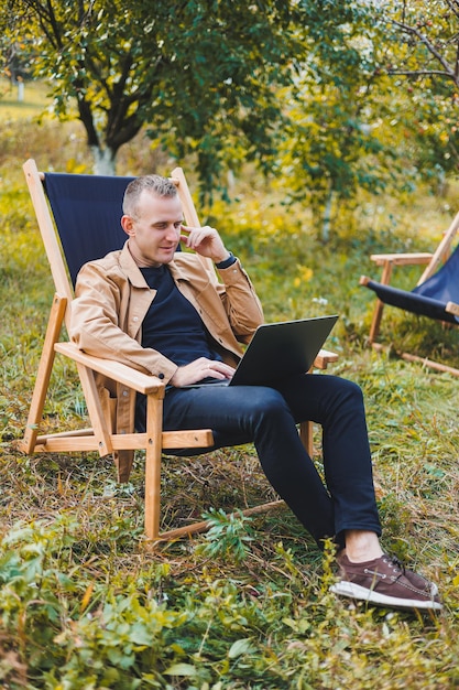 Freelancer man with laptop working in garden sitting in wooden chair outdoors A young blogger works on a computer in a public park Place of work remote work