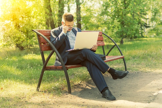 Freelancer programmer sitting at laptop on a bench in Park outdoors businessman working on his laptop connect to wireless internet in rural area Man with laptop at summer park on bright day