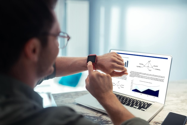 Freelancer wearing glasses reading message on smart watch sitting in front of laptop