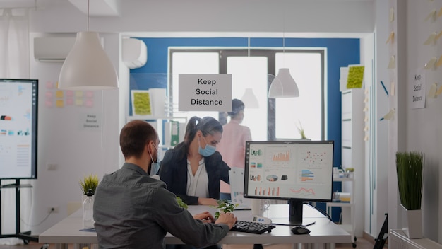 Freelancer with face mask working at financial graphics on pc sitting in business office. Coworkers talking in background about marketing mantain social distancing to prevent infection with covid19