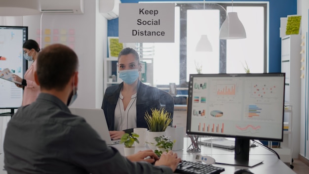 Freelancer with face mask working at financial graphics on pc sitting in business office. Coworkers talking in background about marketing mantain social distancing to prevent infection with covid19